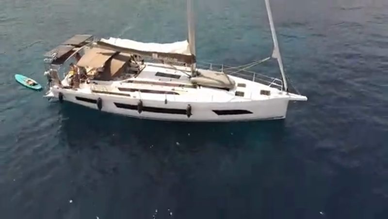 Drone film flying over a busy turquoise anchorage and approaching a charter yacht with guests aboard