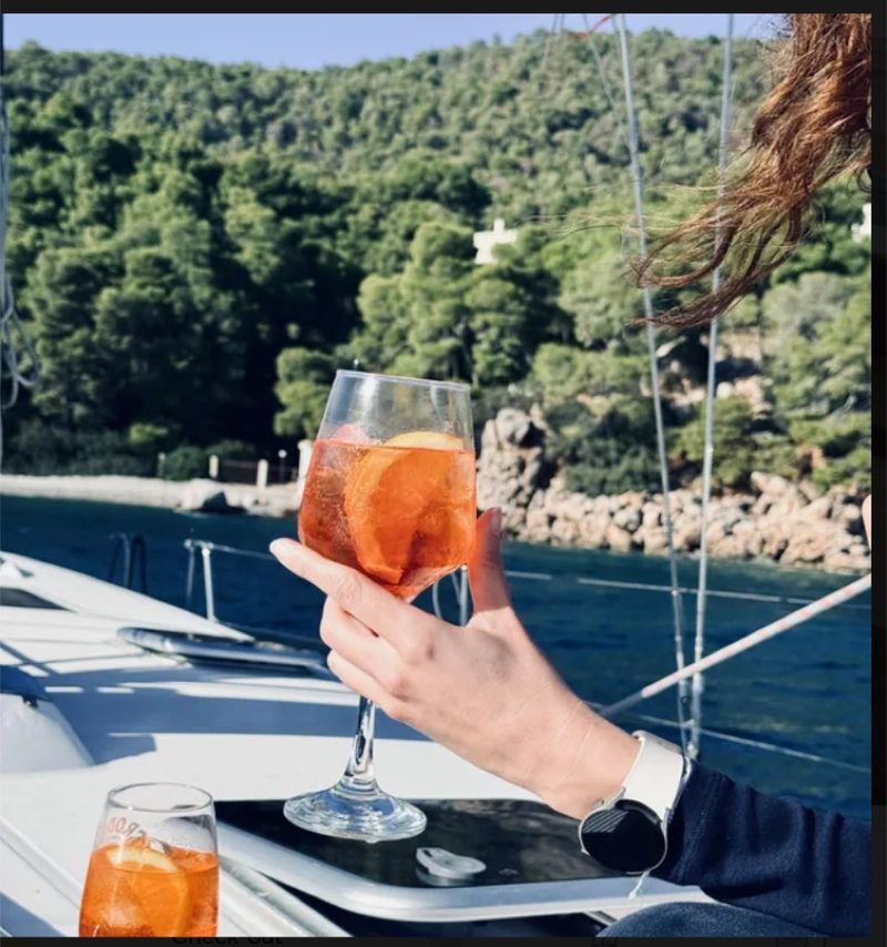 A guest holding an Aperol spritz on the deck of a charter yacht in a green Ionian bay