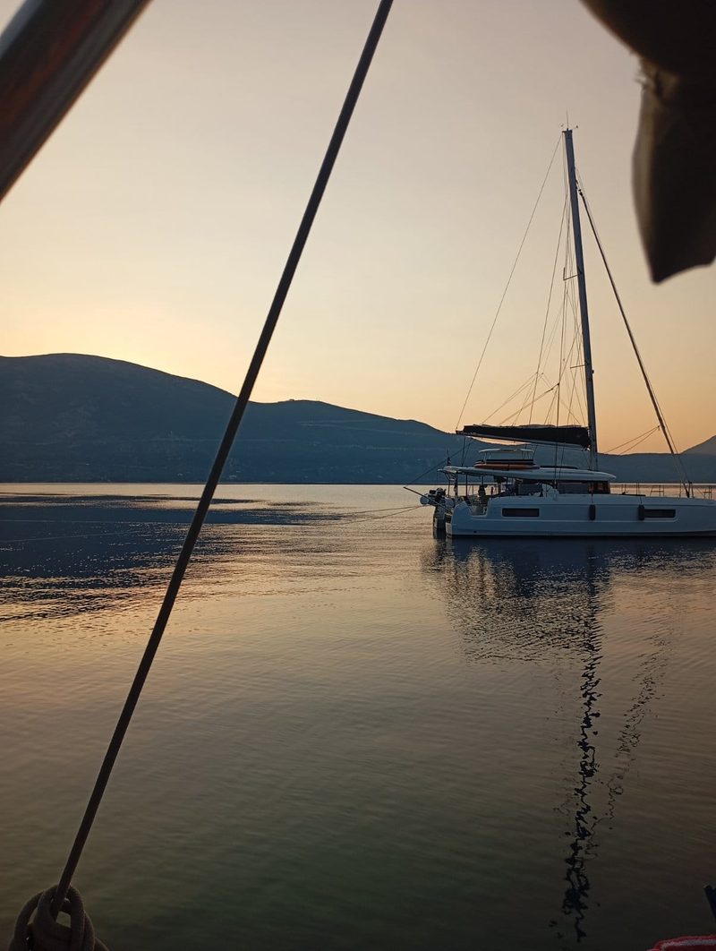 View under the boom of a yacht towards a catamaran silhouetted on a mirror-calm sea at dusk