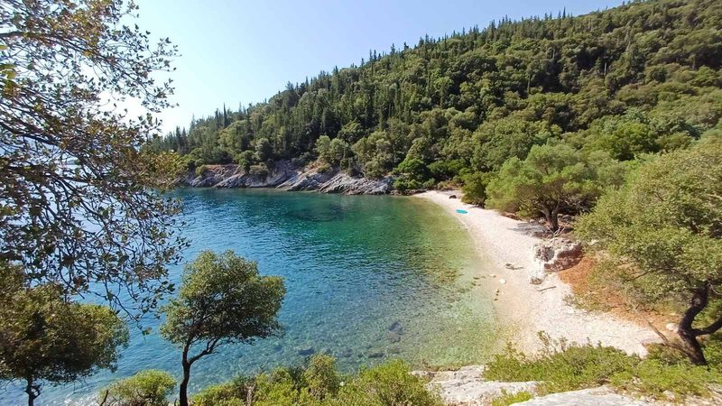 Empty white pebble beach and pine-covered headland beside a clear Ionian bay reached by sailing yacht