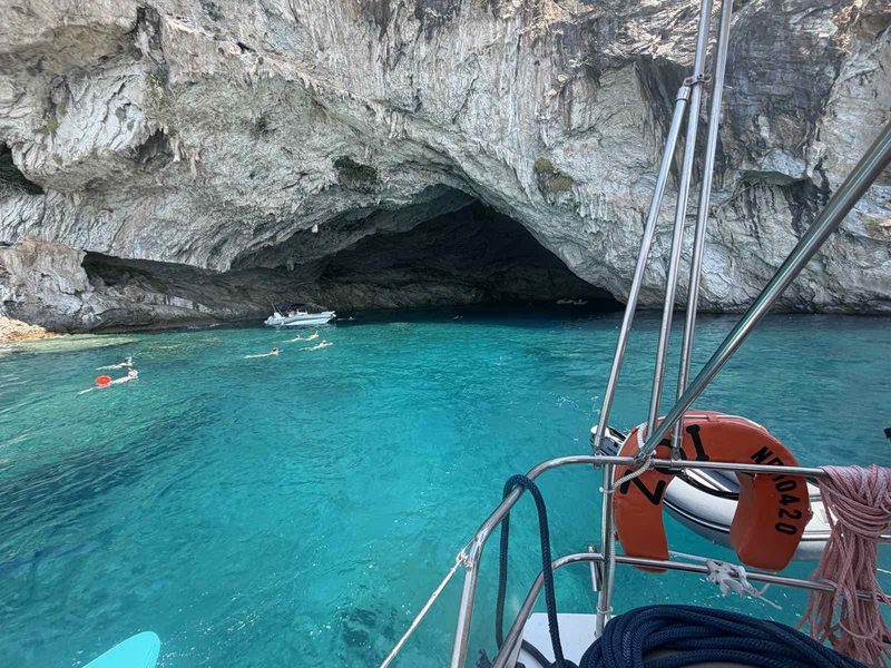 View from the bow of a charter yacht towards a limestone sea cave over bright turquoise water in the Ionian
